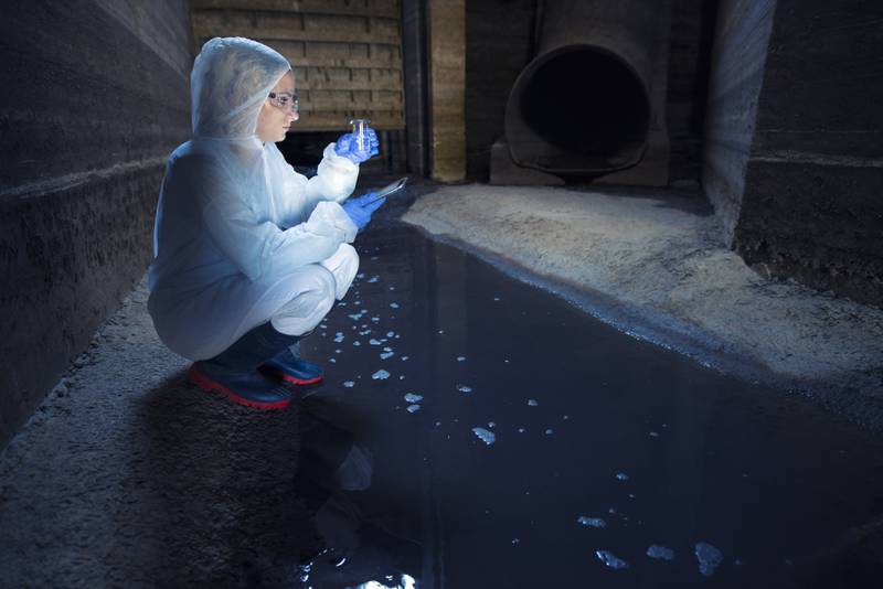 Mulher com roupas para proteção e tubo de ensaio medindo qualidade de água do esgoto