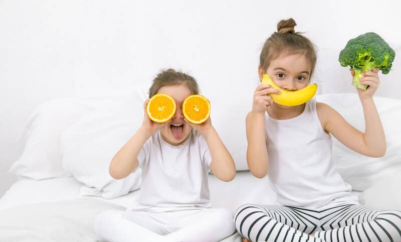 10 alimentos que contribuem para uma boa noite de sono nas crianças; duas meninas de roupas brancas sentadas sobre cama brincam com uma banana e laranja sobre olhos e boca