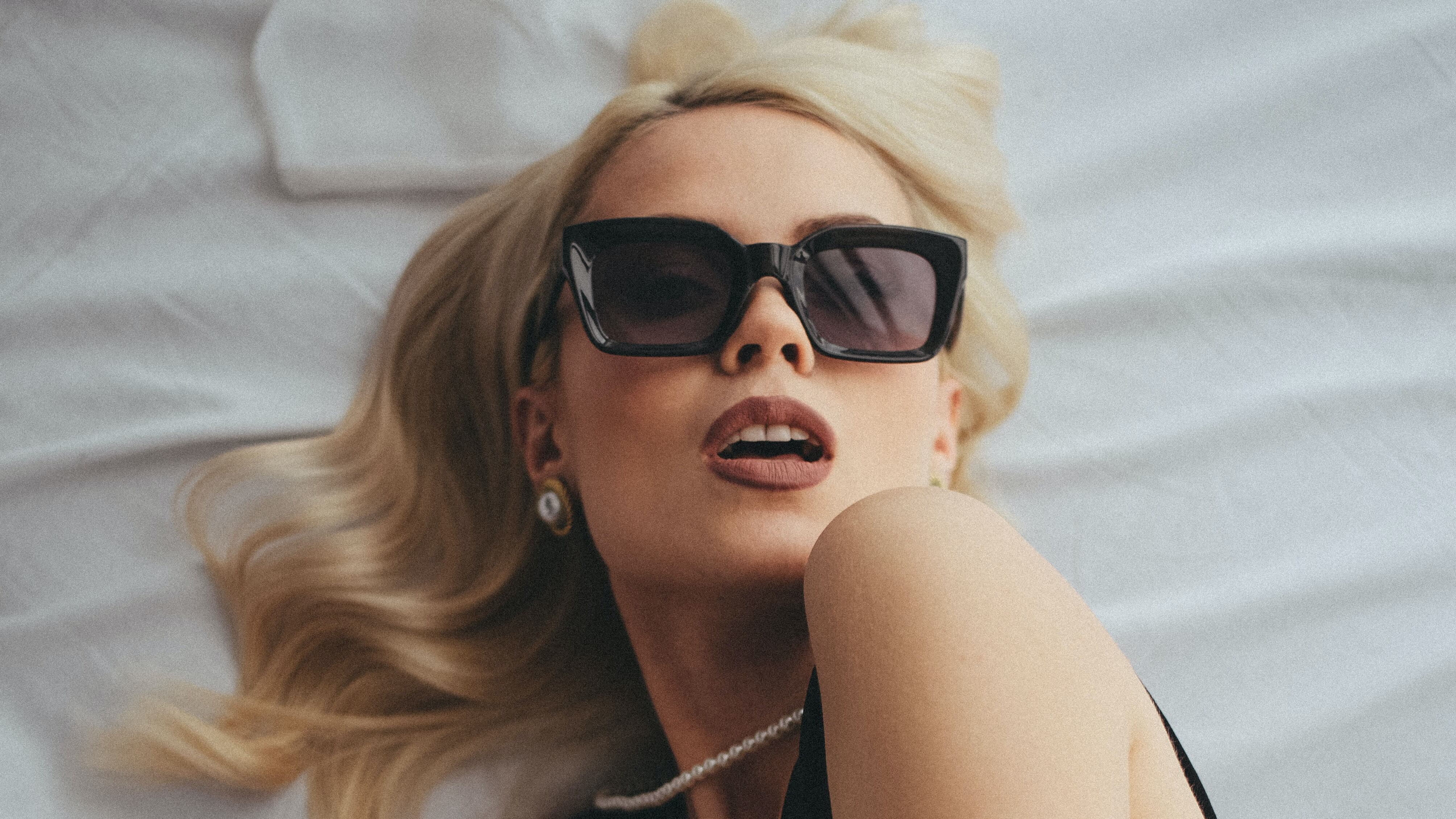 Mulher branca e loira usando óculos escuros e colar de pérolas deitada ao lado de frasco de perfume