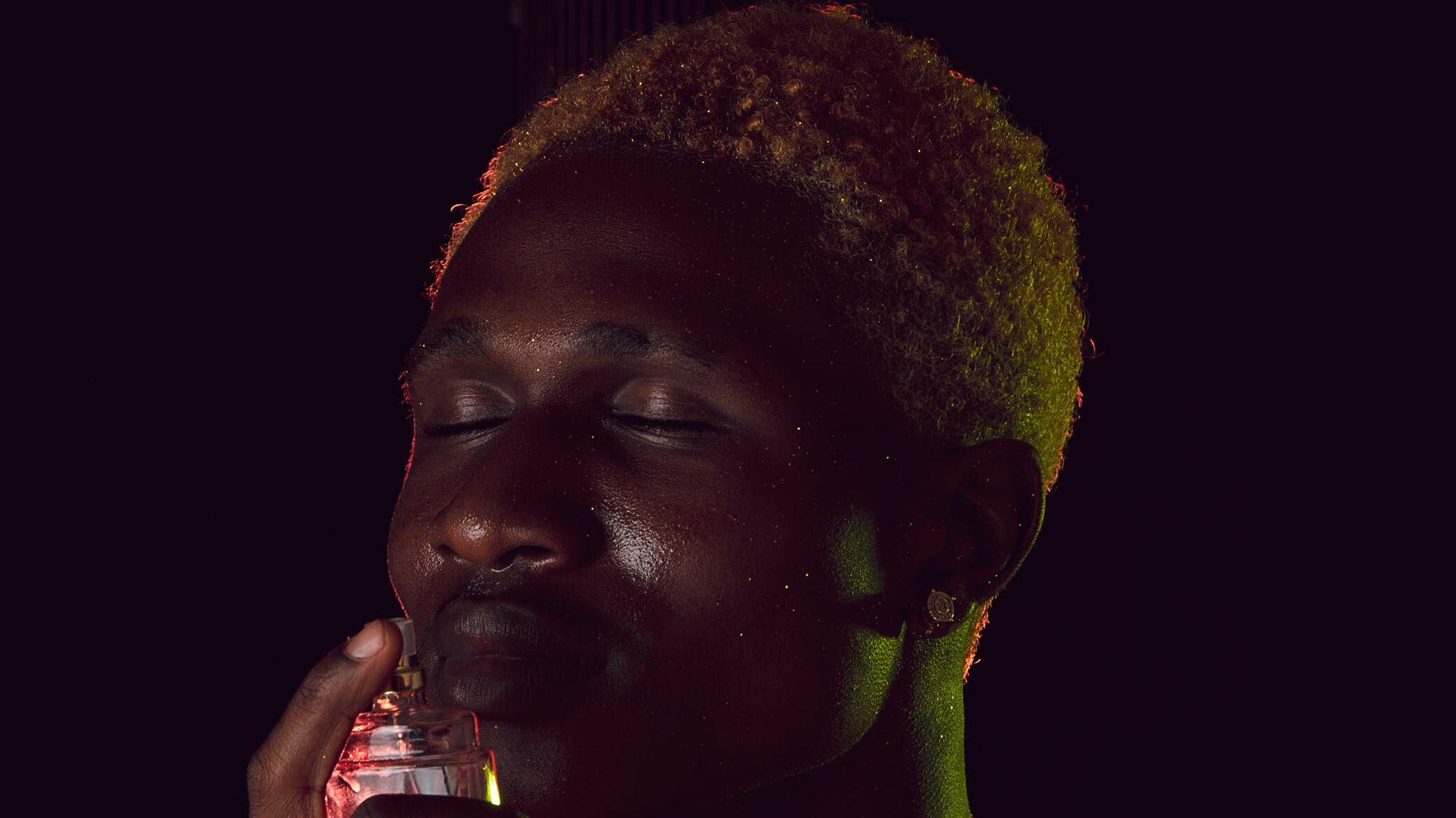 Homem negro com cabelos loiros sentindo cheiro de frasco de perfume vermelho