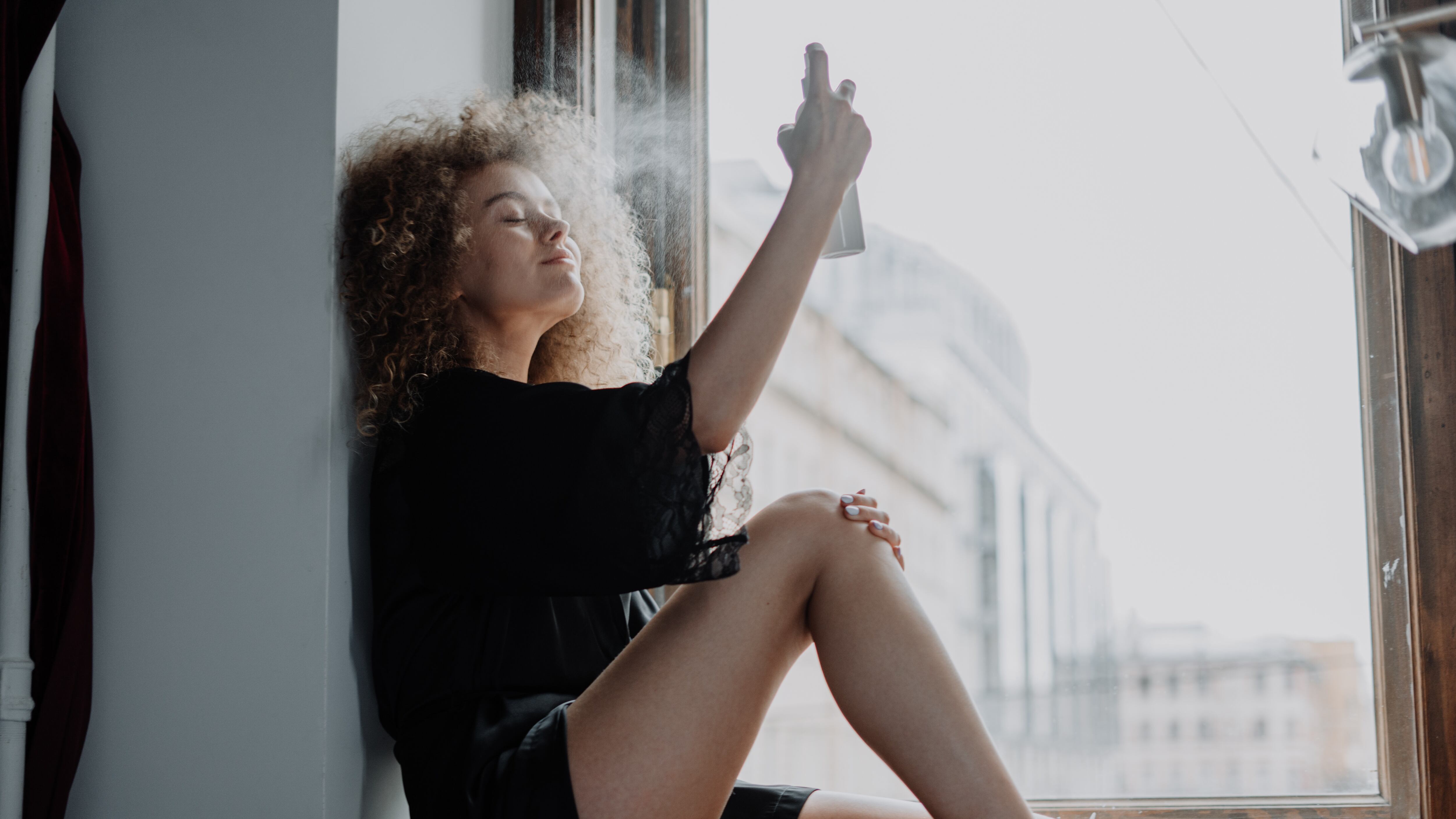 Mulher de cabelo cacheado sentada na janela espirrando body splash no rosto