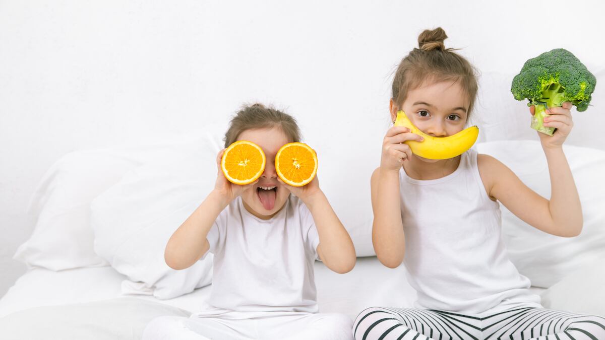 10 alimentos que contribuem para uma boa noite de sono nas crianças; duas meninas de roupas brancas sentadas sobre cama brincam com uma banana e laranja sobre olhos e boca