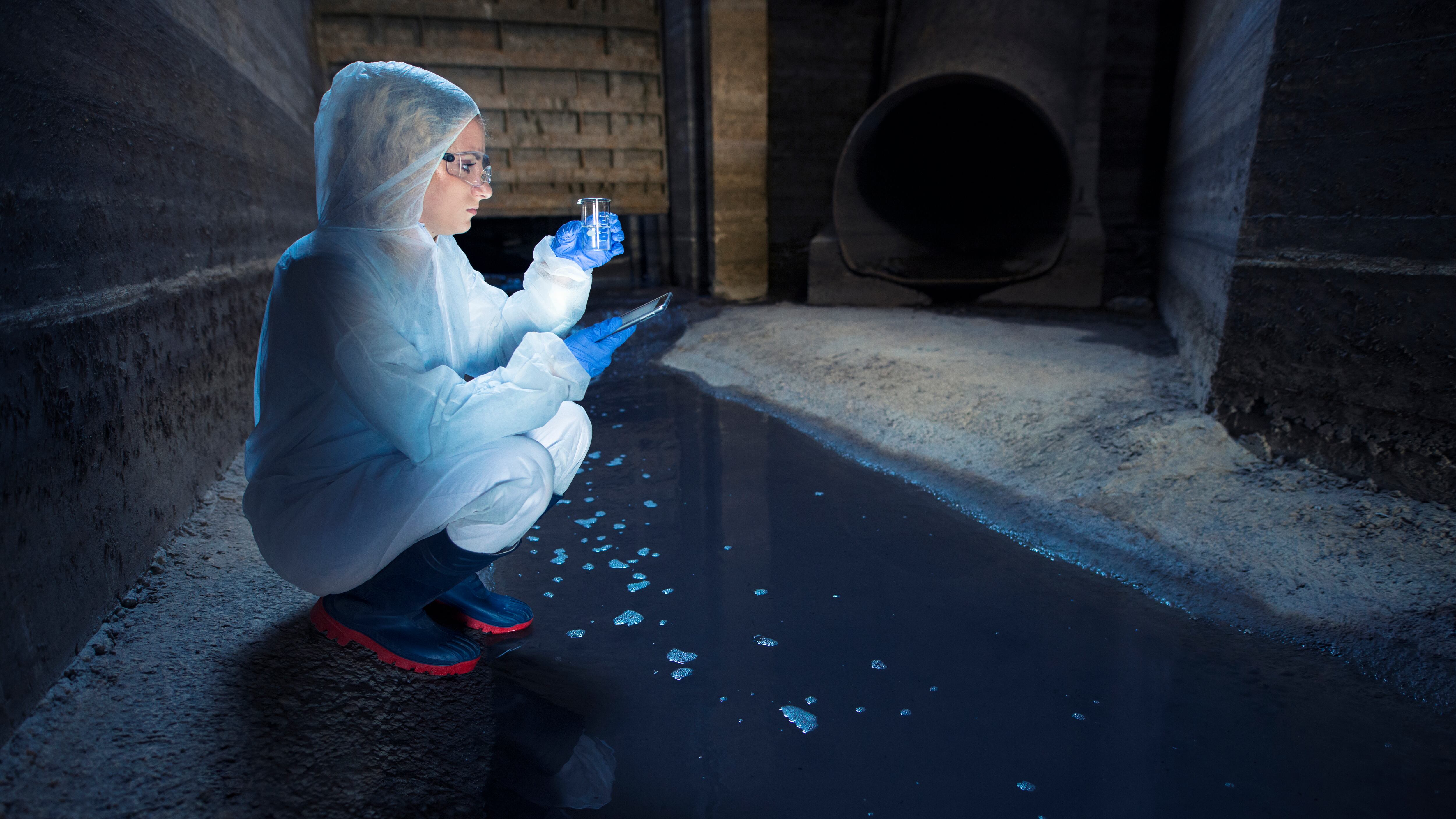 Mulher com roupas para proteção e tubo de ensaio medindo qualidade de água do esgoto