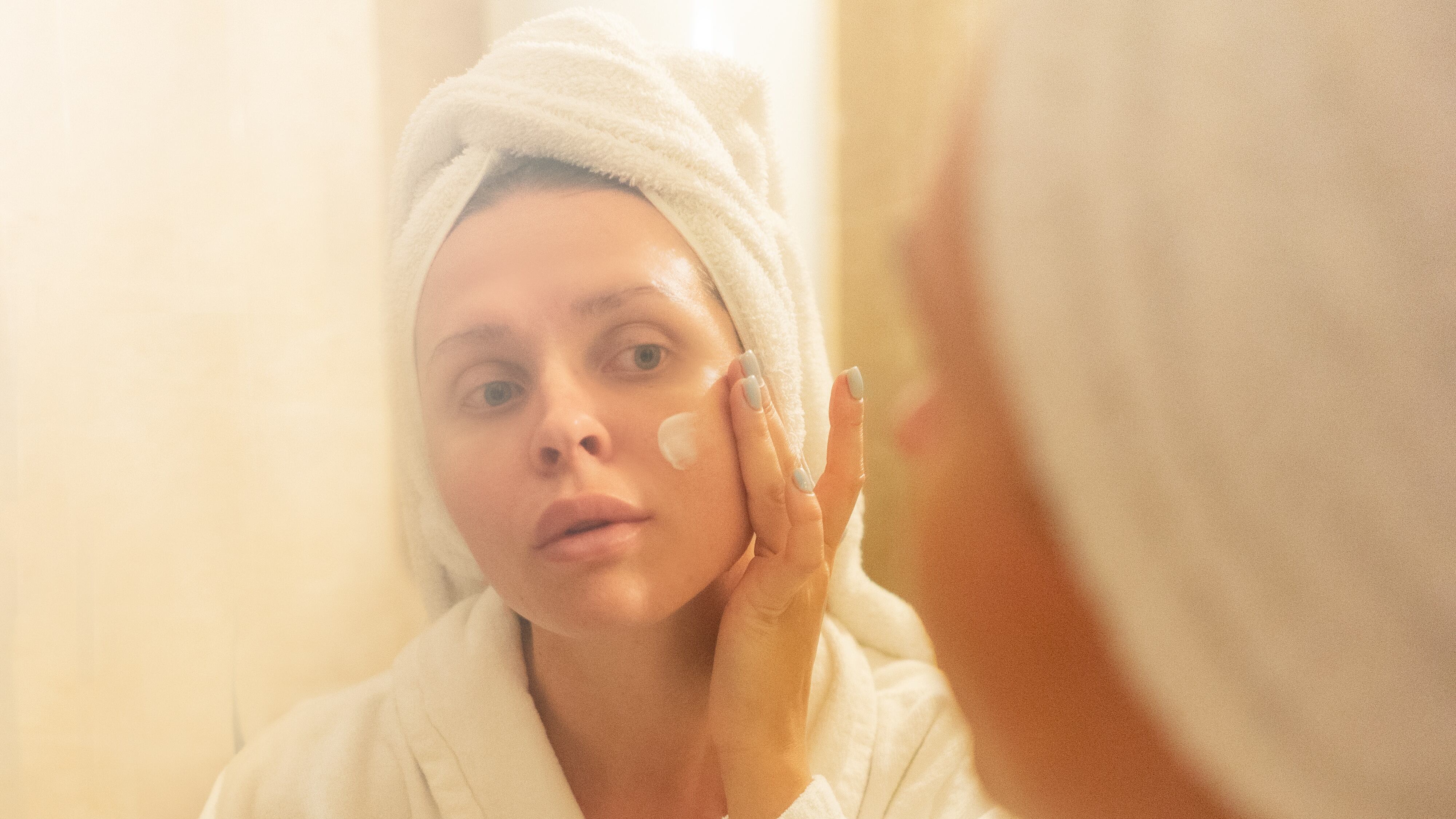 Mulher de roupão e toalha na cabeça passando hidratante no rosto