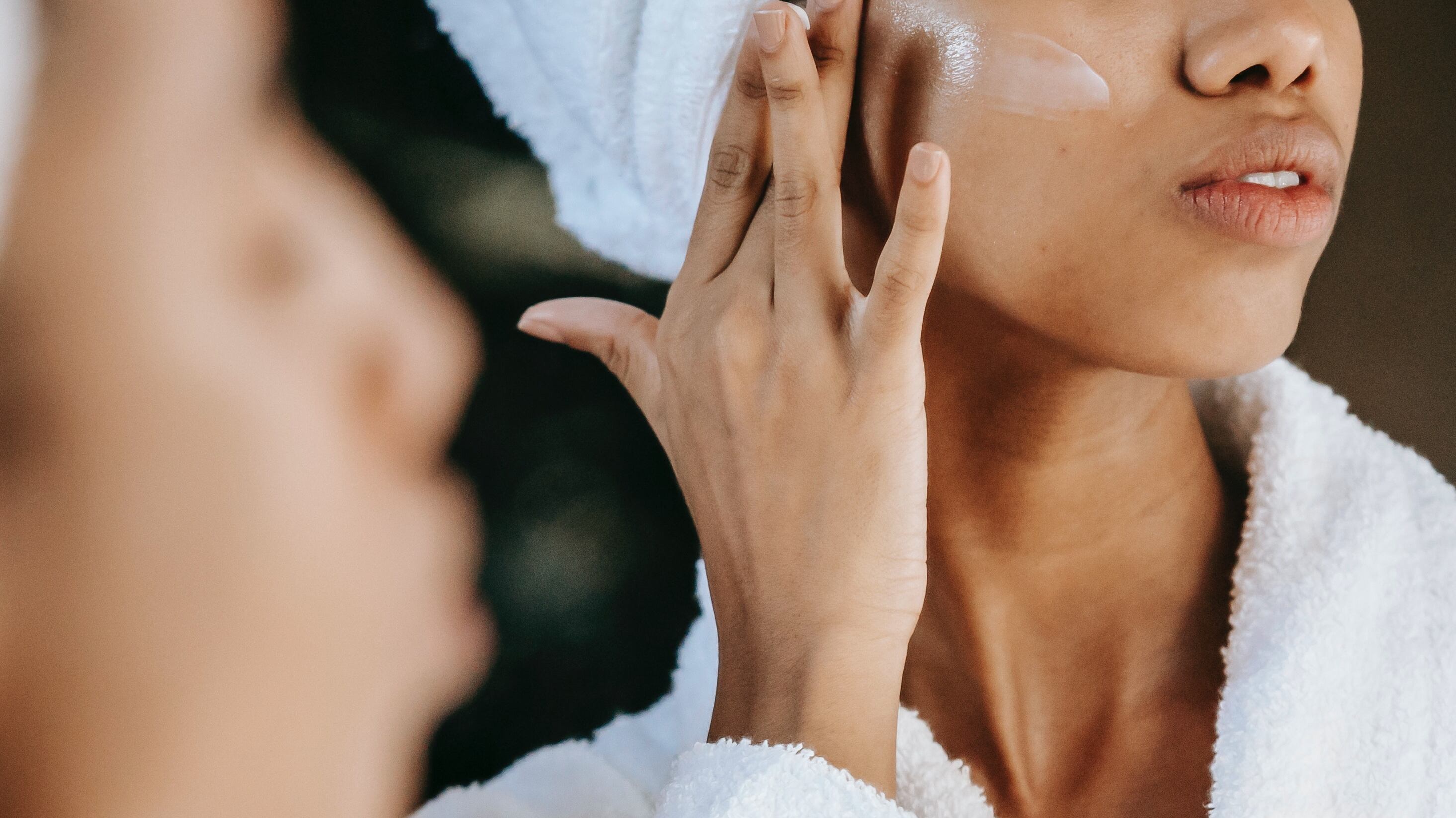 Mulher de roupão e toalha na cabeça em frente ao espelho passando hidratante no rosto