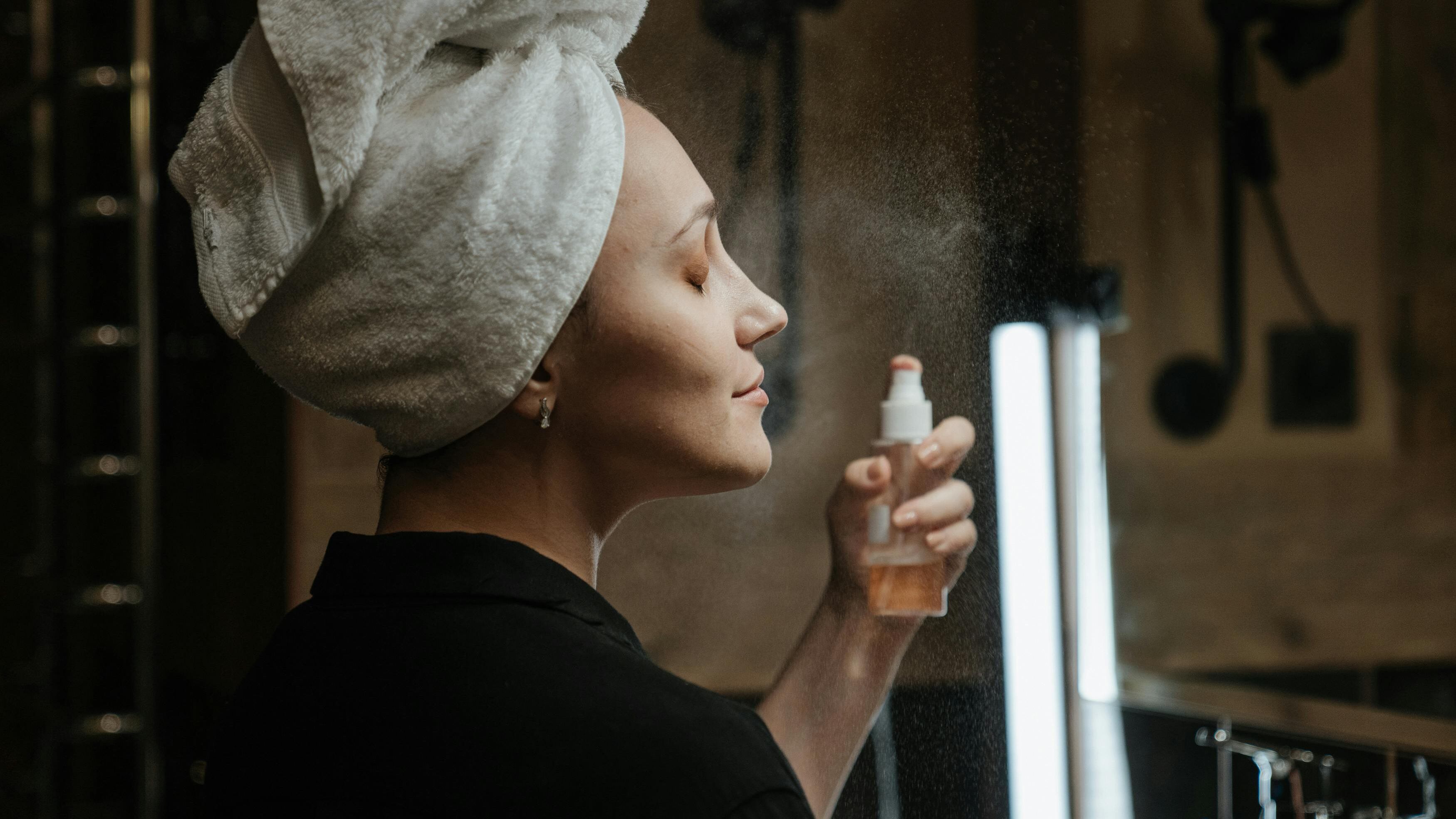 Mulher com toalha na cabeça espirrando spray no rosto