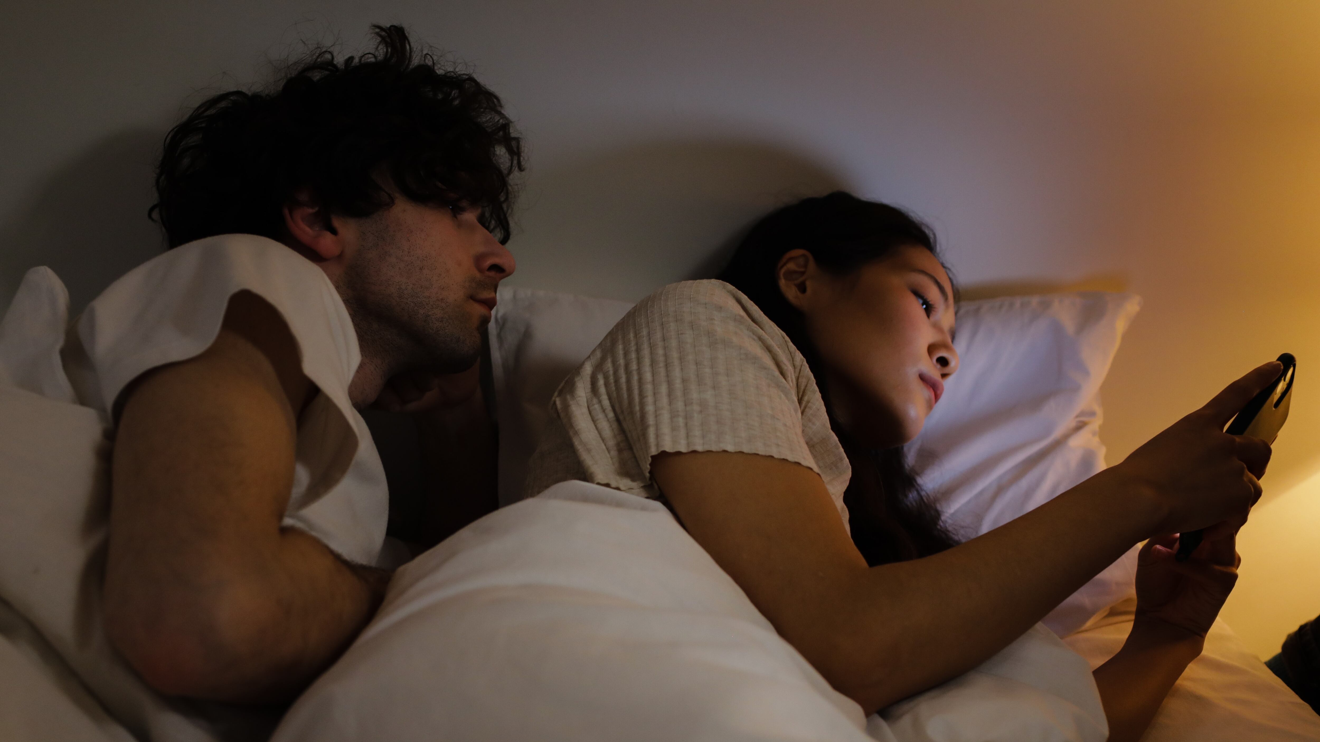 Casal deitado na cama e mulher mexendo no celular com homem bisbilhotando
