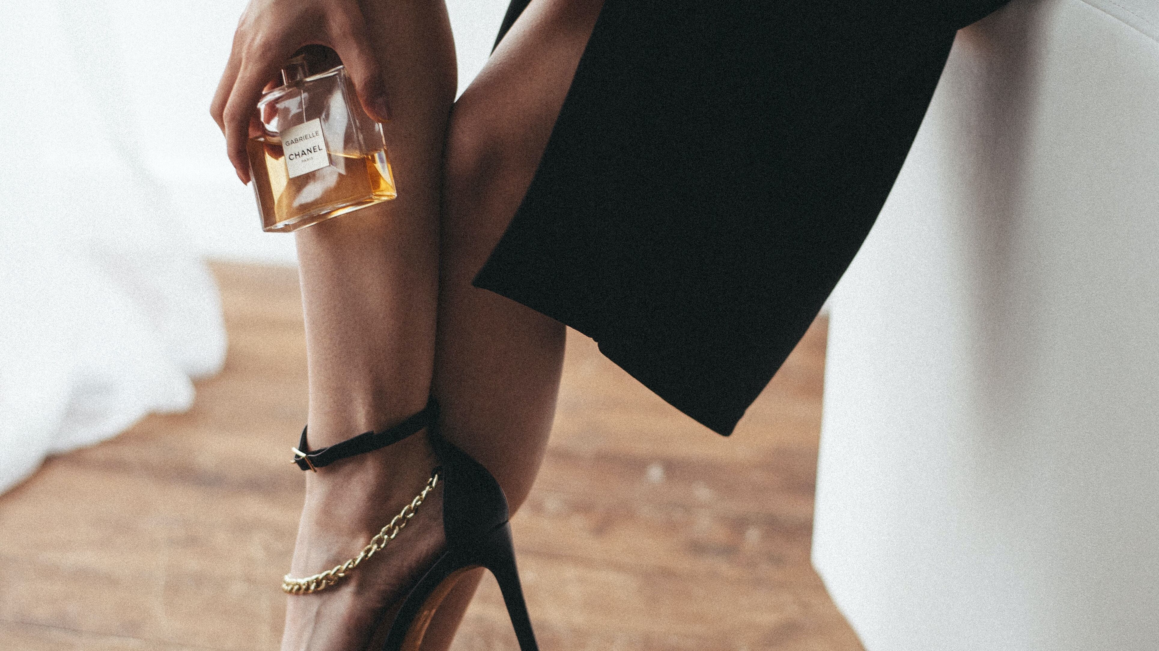 Mulher elegante de vestido preto e salto fino preto mostrando frasco de perfume Chanel