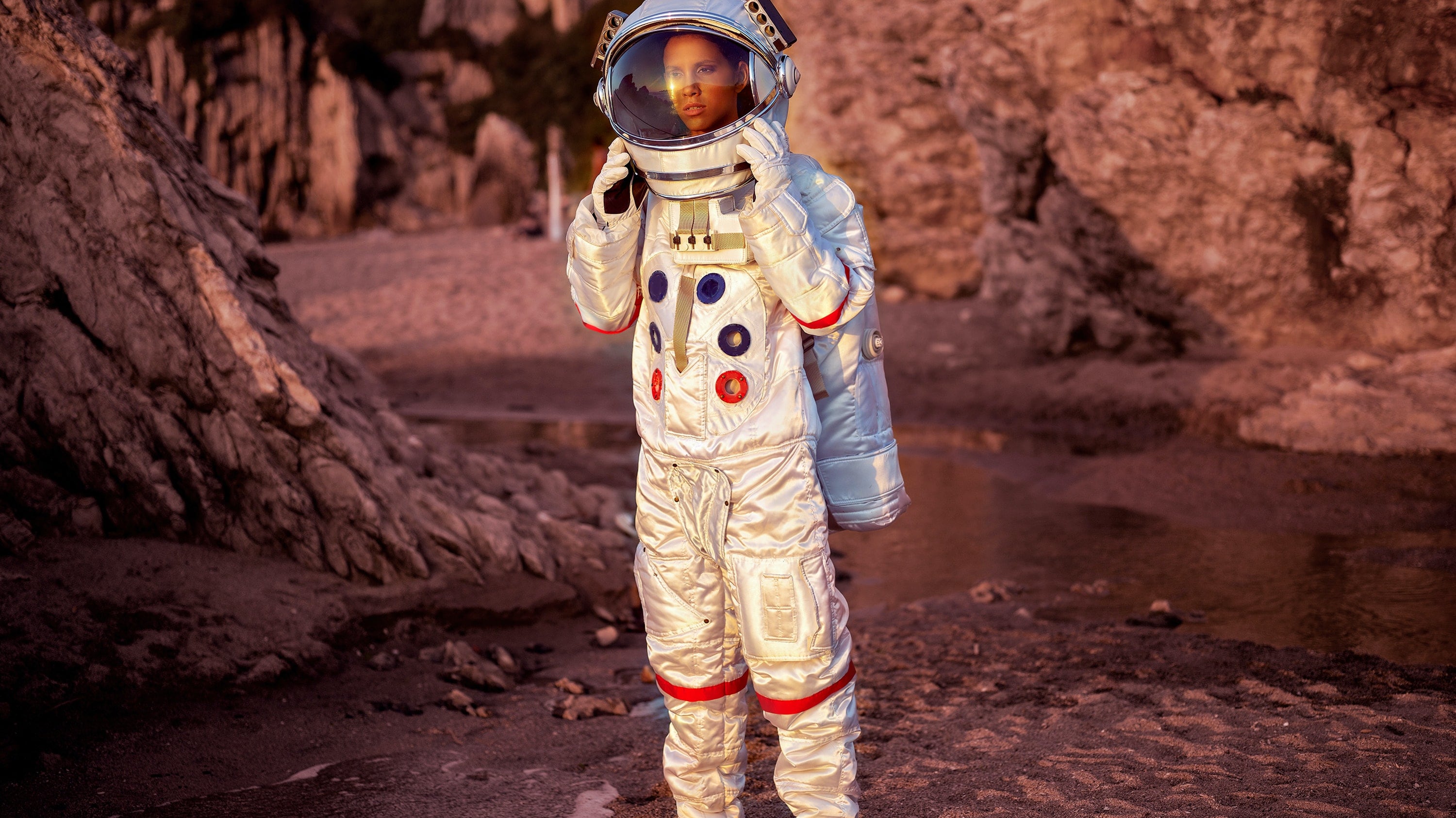 Mulher vestida de astronauta próxima à pedras vermelhas e corredeira de água ao fundo