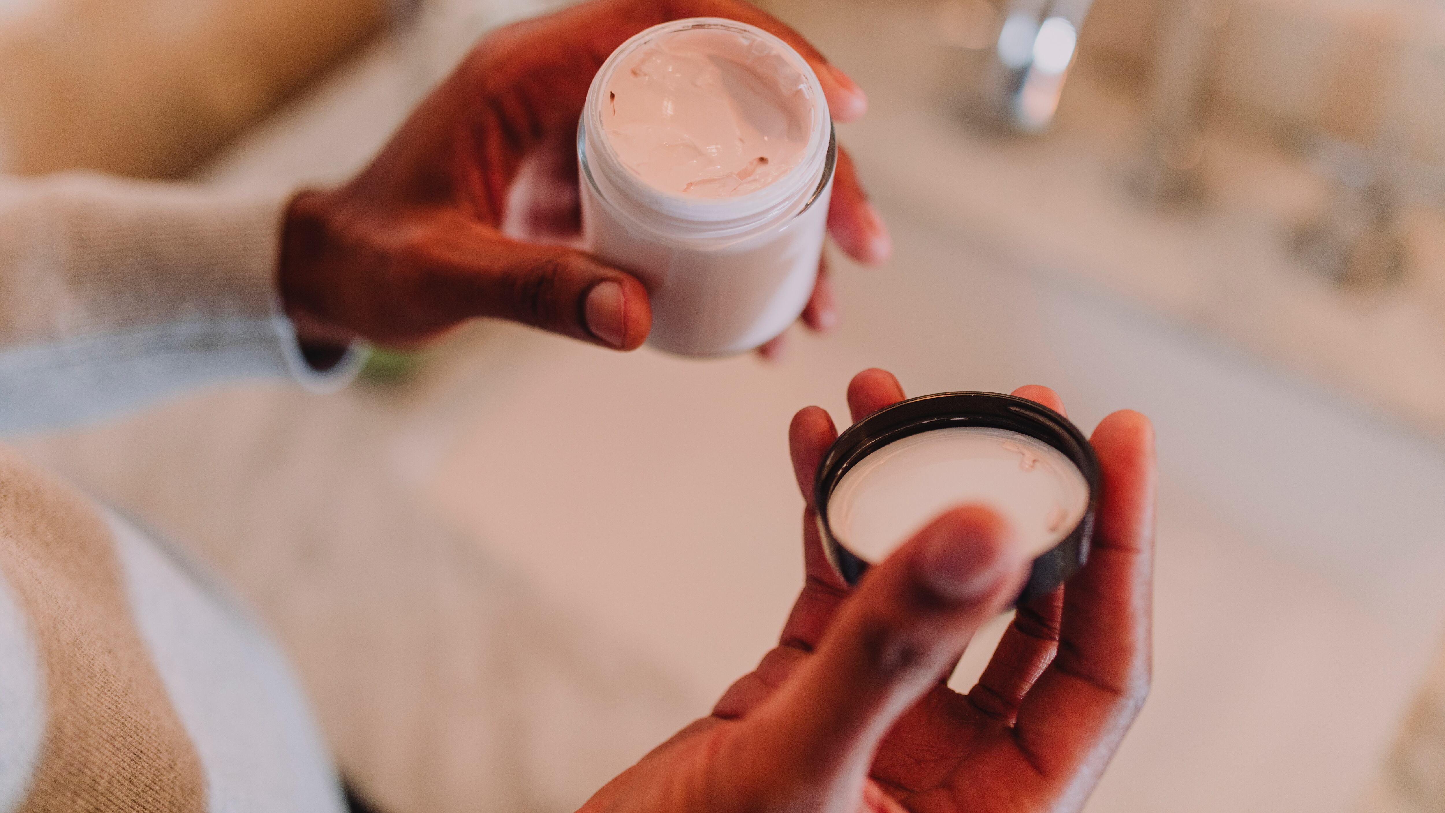 Mãos negras segurando pote hidratante em frente à pia do banheiro