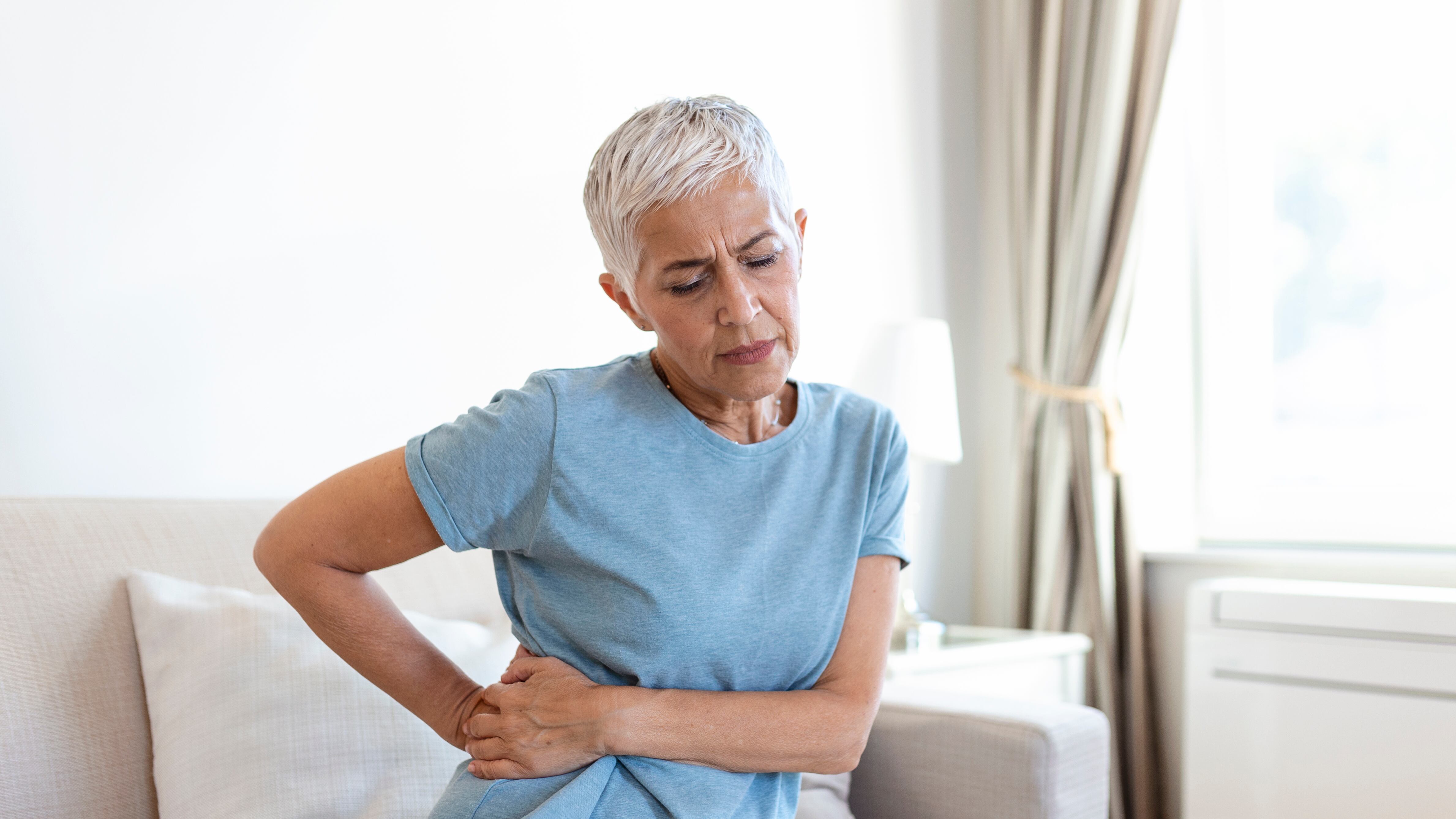 Senhora de cabelo branco curto e camisa azul sentada e colocando as mãos nos rins