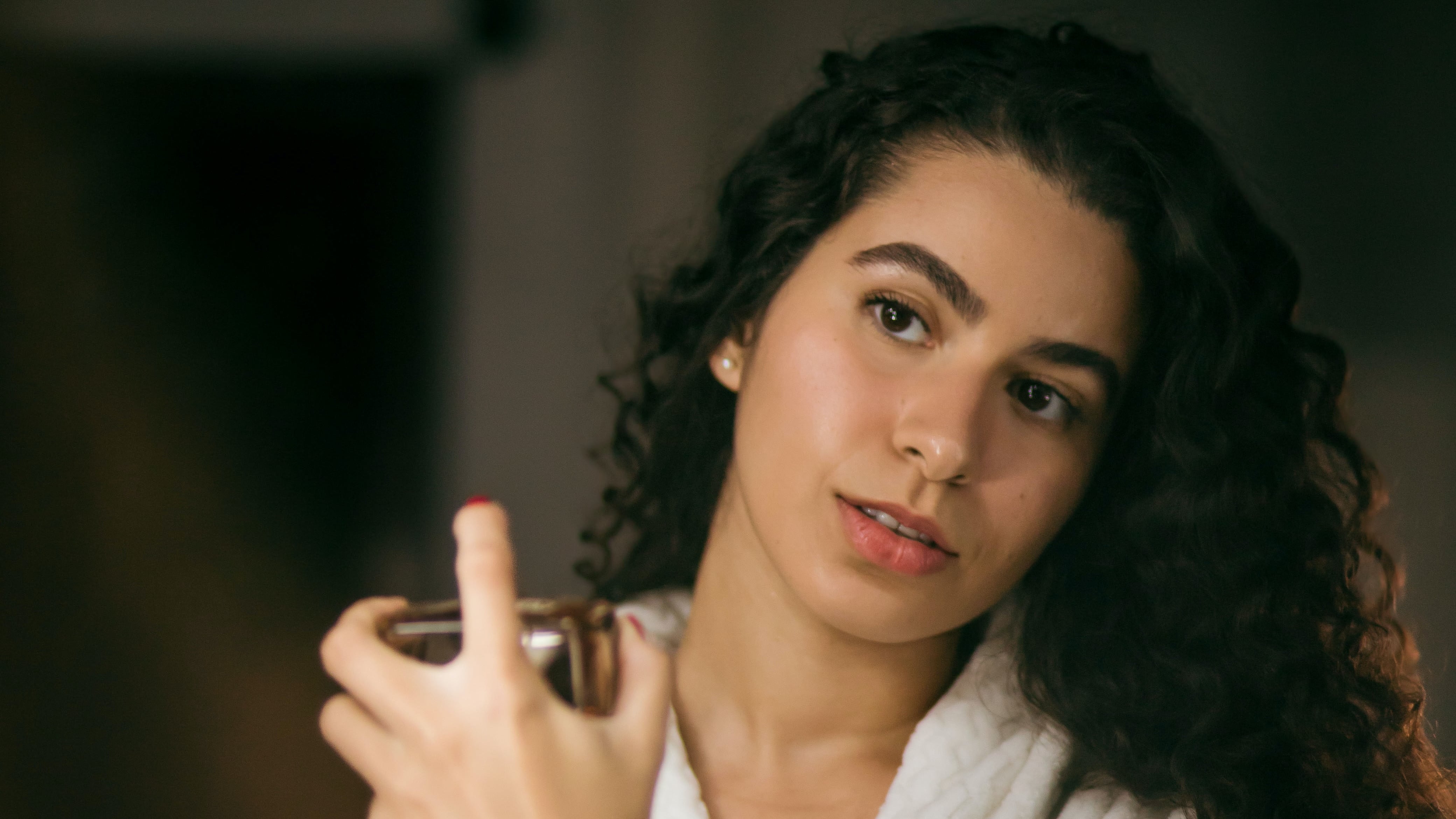 Mulher branca de roupão em frente ao espelho com cabelo cacheado preto e batom vermelho borrifando perfume