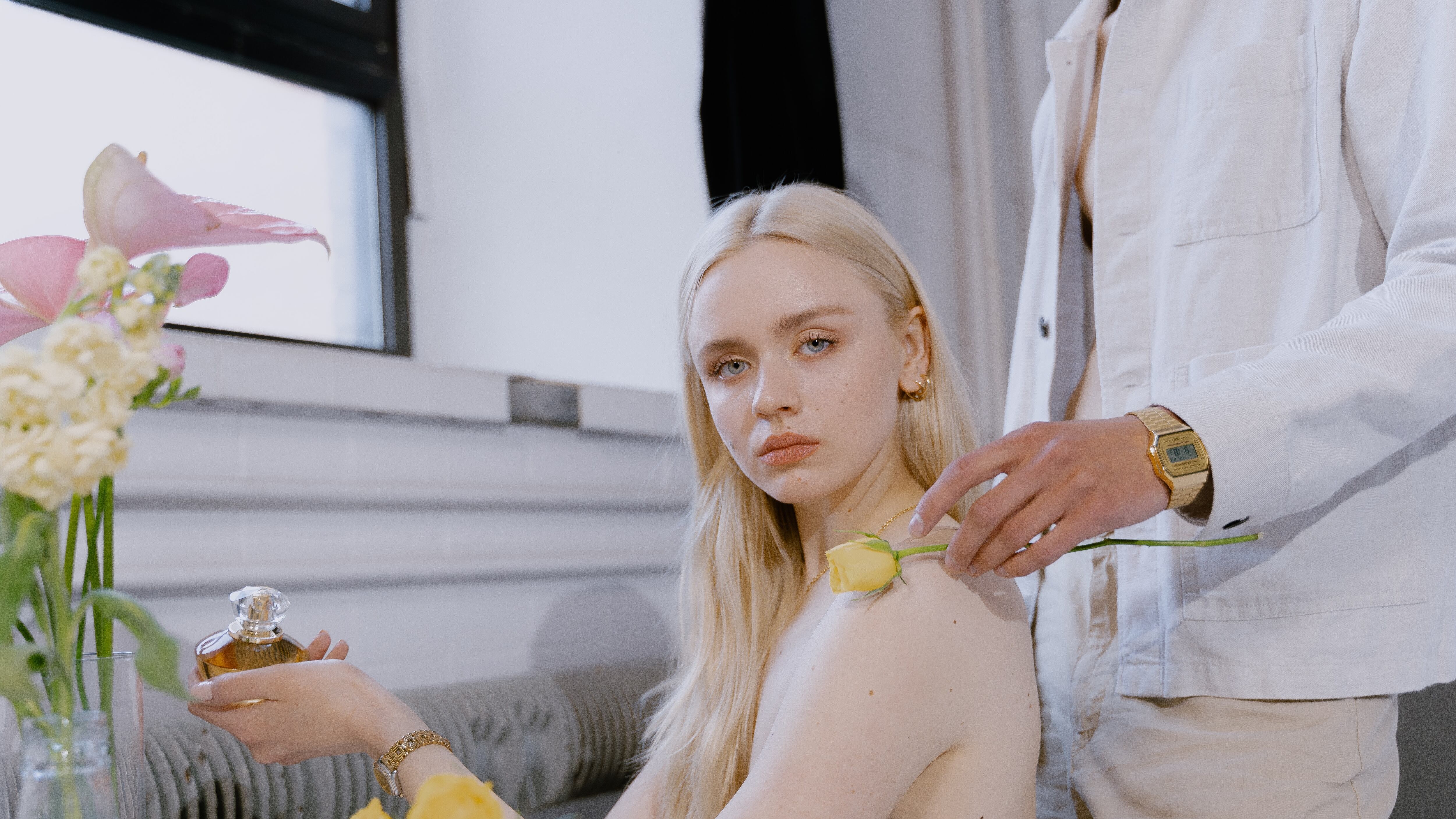 Mulher branca e loira segurando frasco de perfume com homem colocando uma flor amarela em seu ombro