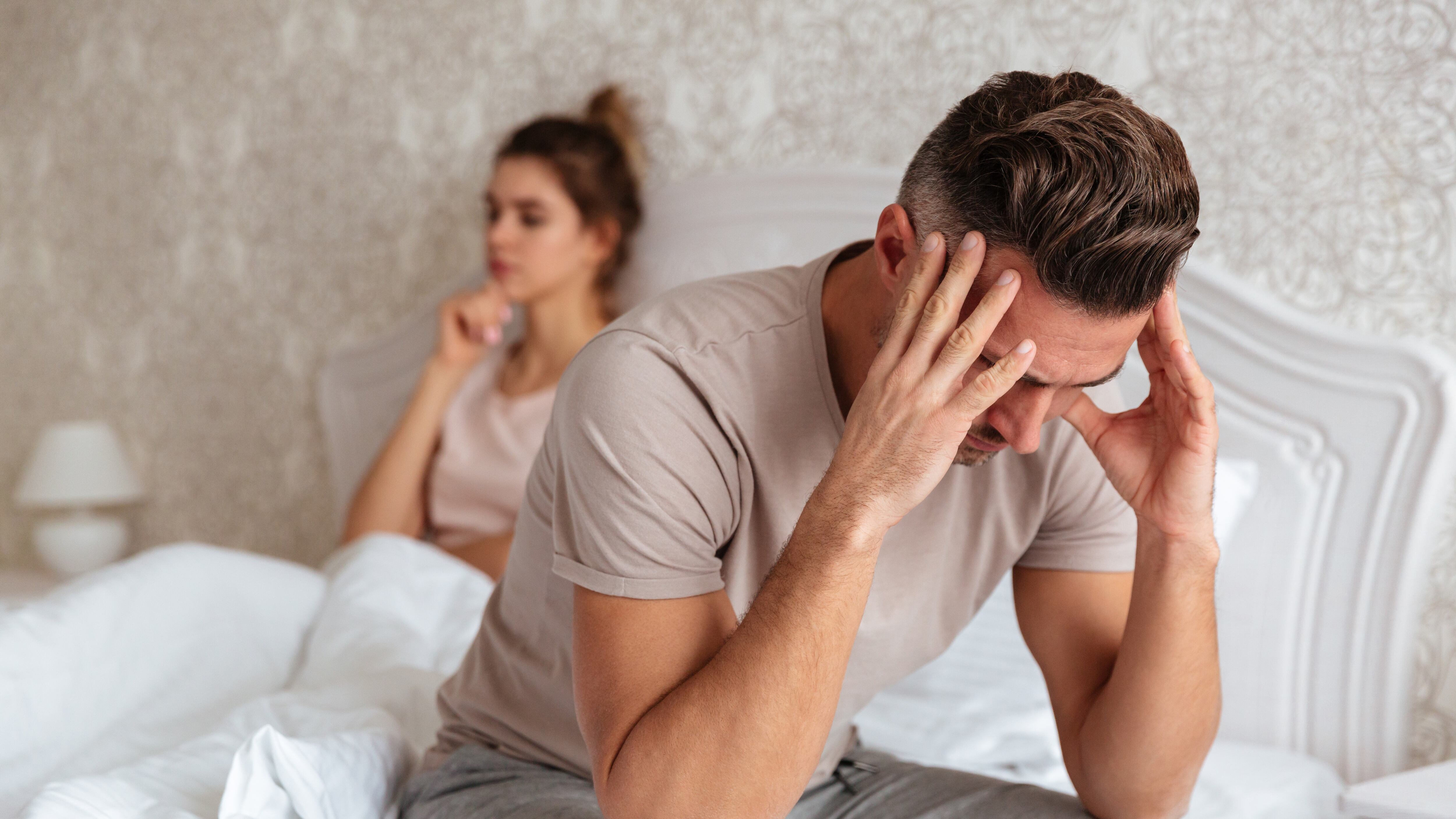 Homem cabisbaixo sentado na beira da cama com mulher ao fundo