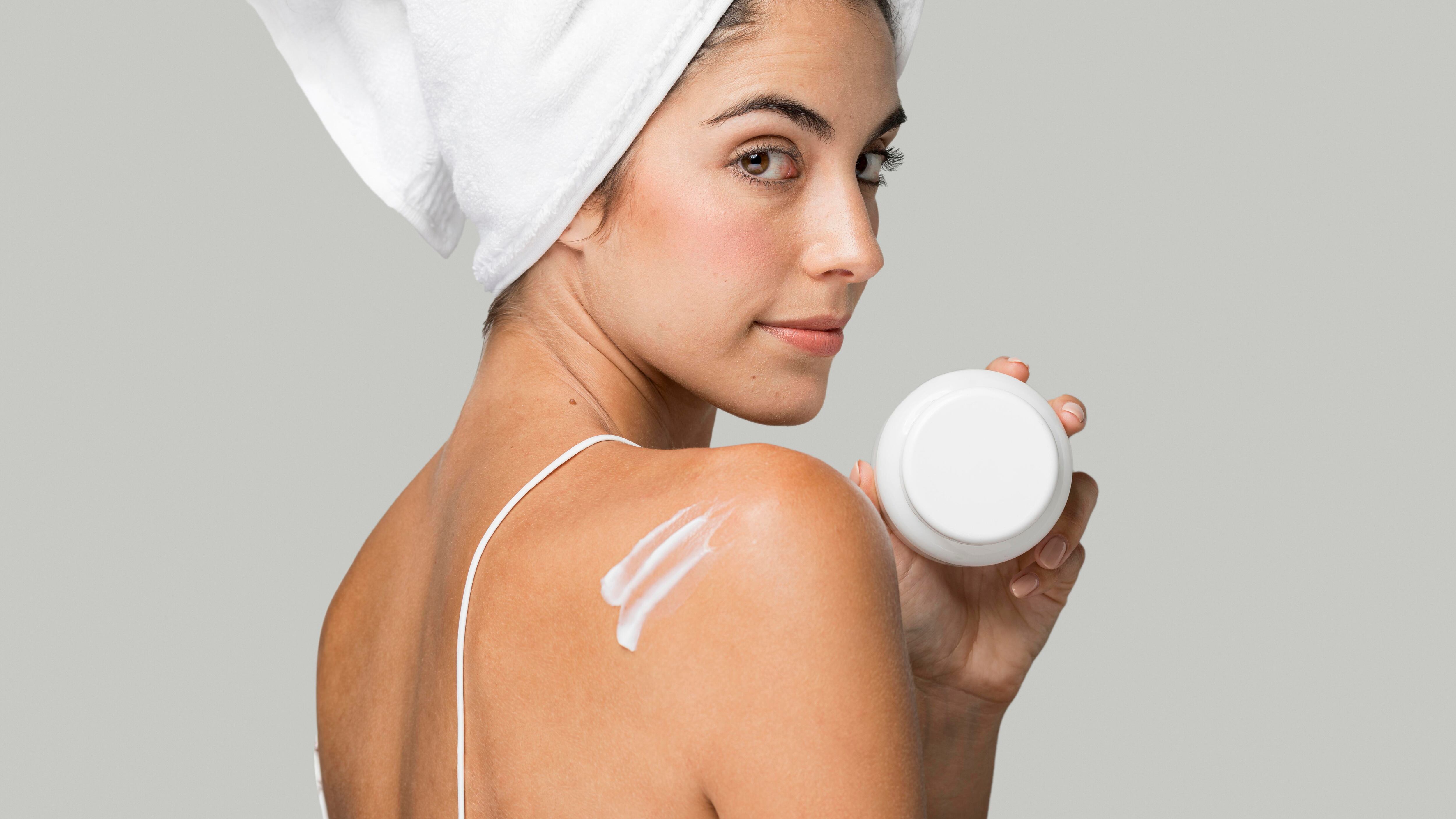 Mulher branca com toalha na cabeça mostrando pote de creme