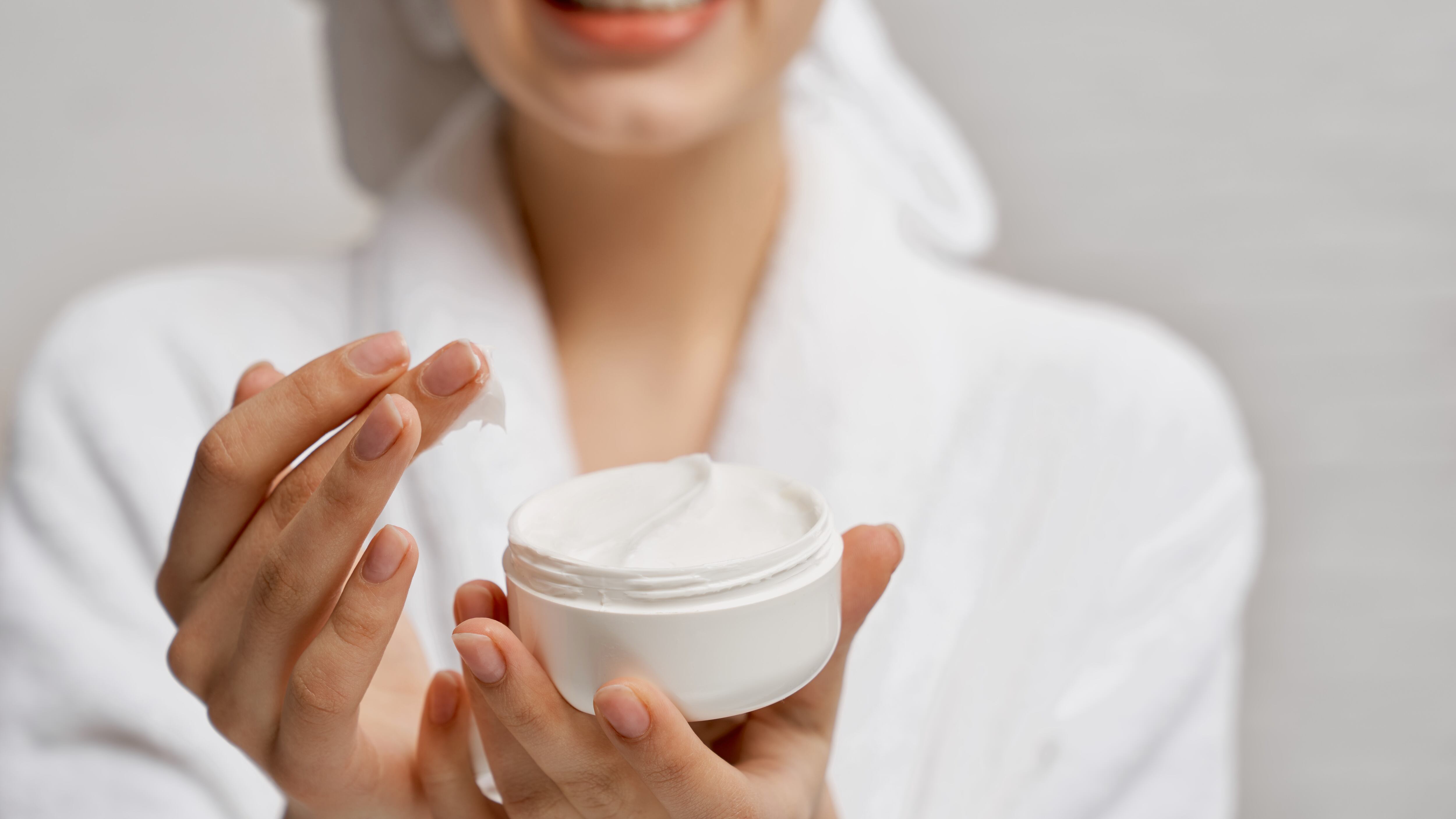 Mulher de roupão e toalha branca na cabeça com pote de creme na mão