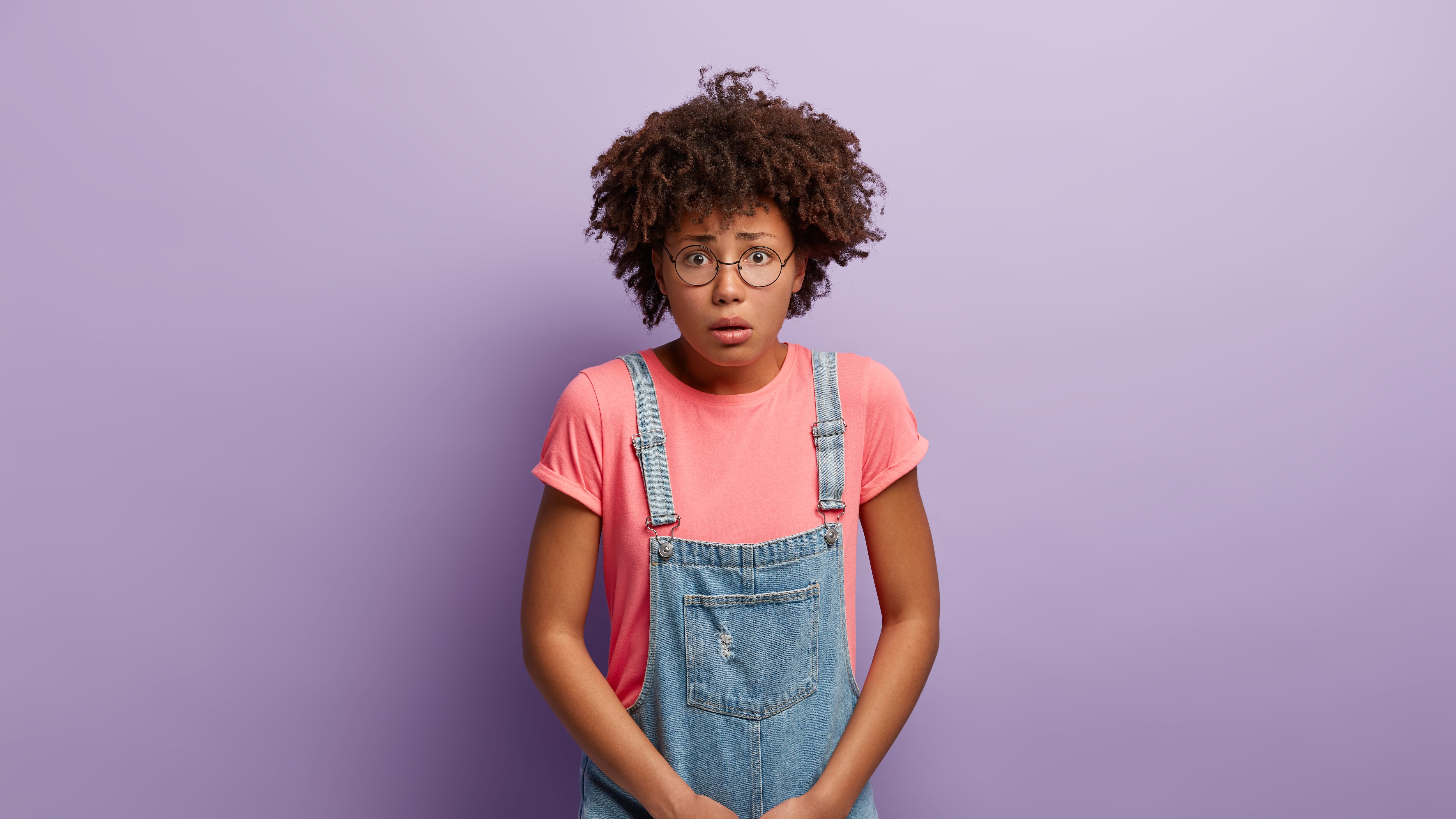 Mulher negra com cabelos crespos vestindo macacão com cara de assustada colocando a mão sob a bexiga